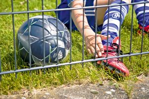 Junge in Sportkleidung verfängt sich mitd em Fußballschuh am Zaun